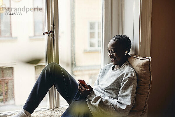 Smiling teenage boy with wireless in-ear headphones using smart phone near window at home