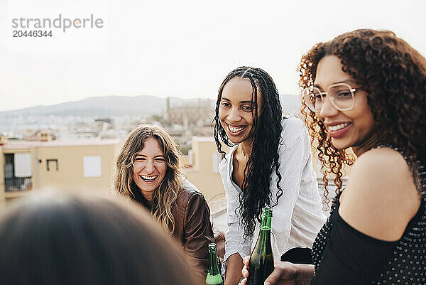 Cheerful female friends holding beer bottles and enjoying at rooftop party