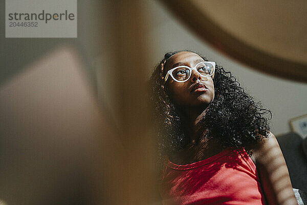 Low angle view of girl with curly hair and wearing eyeglasses looking away at home