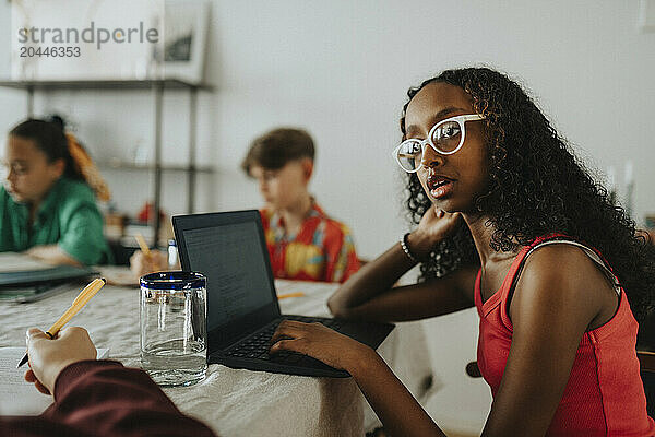 Thoughtful teenage girl using laptop while sitting with friends at home