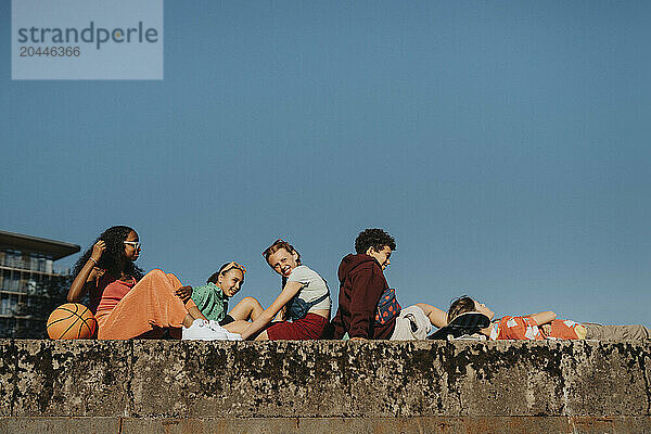 Group of male and female friends sitting on wall under clear sky