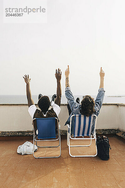 Rear view of male friends sitting on chairs with arms raised on rooftop