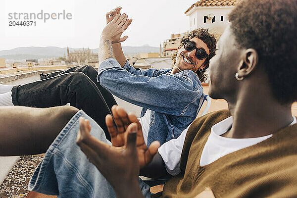 Happy man wearing sunglasses and clapping while sitting with male friend on rooftop