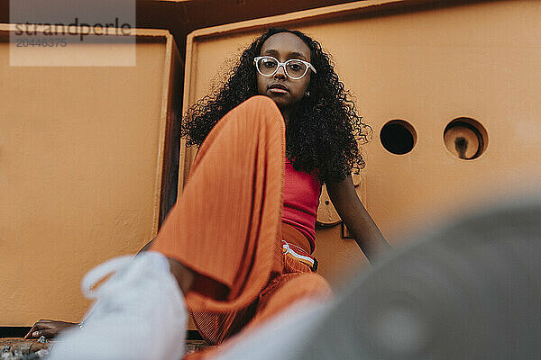 Portrait of girl wearing eyeglasses sitting on ground against metal structure