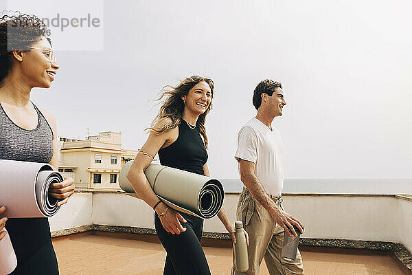 Smiling male and female friends holding yoga mats and walking on rooftop