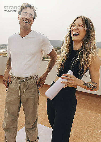 Happy woman holding water bottle and laughing while standing with male friend on rooftop