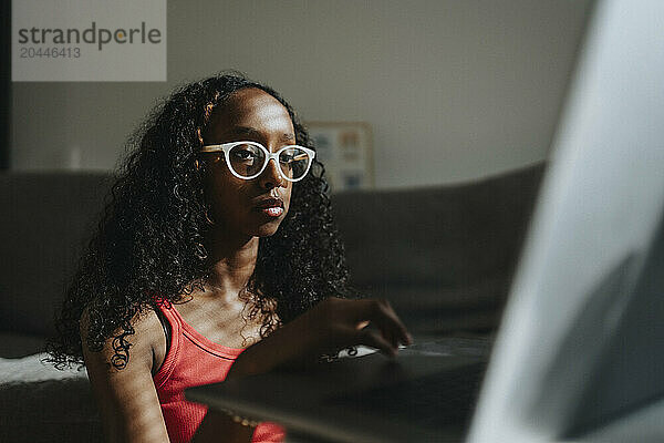 Focused girl with curly hair and eyeglasses studying on laptop at home