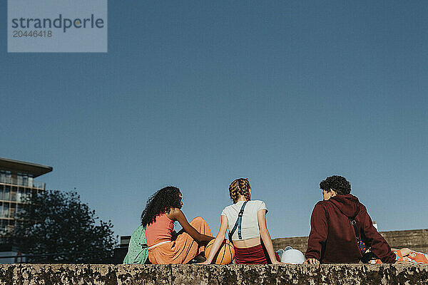 Rear view of friends sitting on wall under blue sky