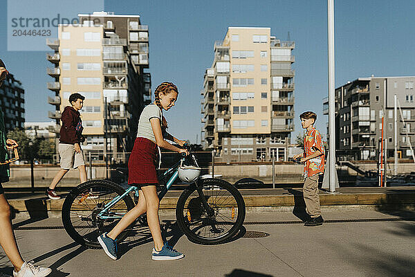 Girl walking with bicycle and friends under blue sky at street