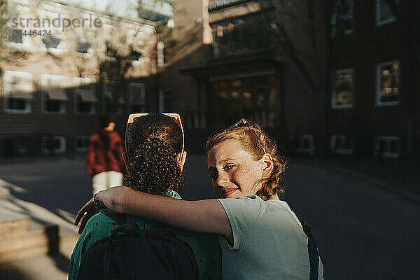 Portrait of smiling girl with arm around friend looking over shoulder while walking at school campus