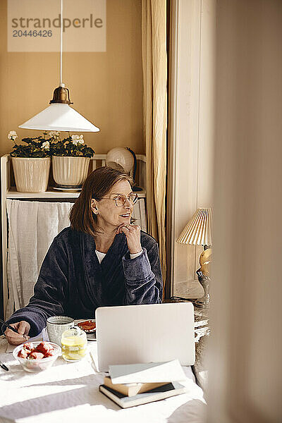 Senior woman with hand on chin sitting at dining table in home