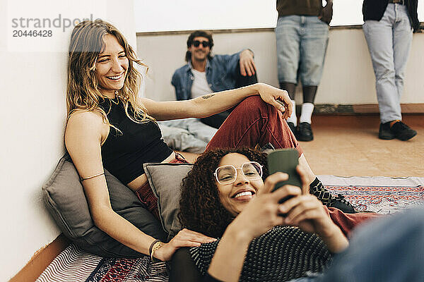 Smiling woman using smart phone lying down near female friend sitting on rooftop