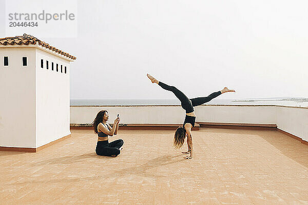 Woman photographing female friend doing handstand on building rooftop against sky