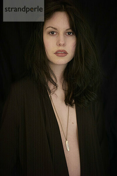 A young woman with disheveled dark hair and a neutral expression is wearing a dark open cardigan and a necklace with a white pendant. The background is black  highlighting her presence.