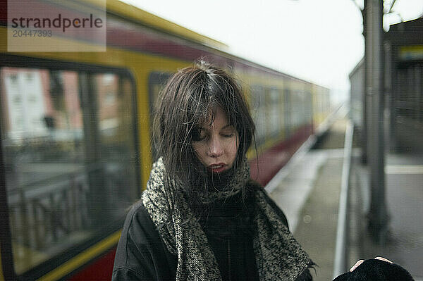 A woman stands on a train platform  her face partially obscured by windswept hair  as she appears deep in thought.