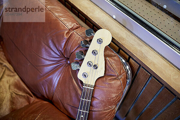 An electric bass guitar rests horizontally across a worn brown leather couch  its headstock in focus against a blurred indoor background.