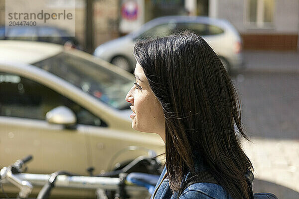 A woman with long dark hair is photographed in profile  looking ahead on a sunny day  with cars and bicycles in the background.