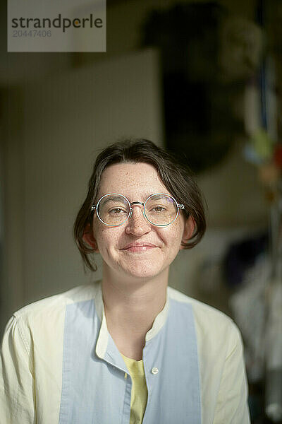 A smiling young woman with short hair wearing round glasses and a light blue shirt over a yellow top is standing indoors  with a blurred background suggesting a room with objects.