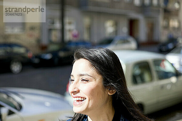 A smiling woman outdoors on a sunny day  with blurred street and parked cars in the background.