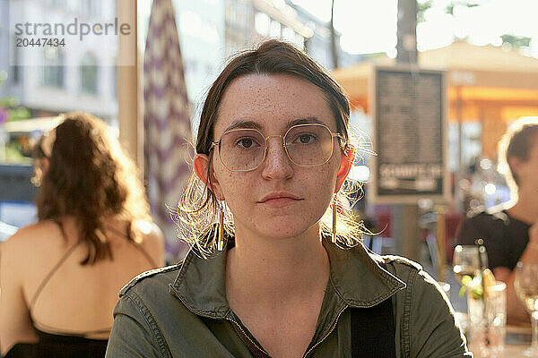 A young woman with glasses is looking directly at the camera with a serious expression  with blurred people and urban street life visible in the background.