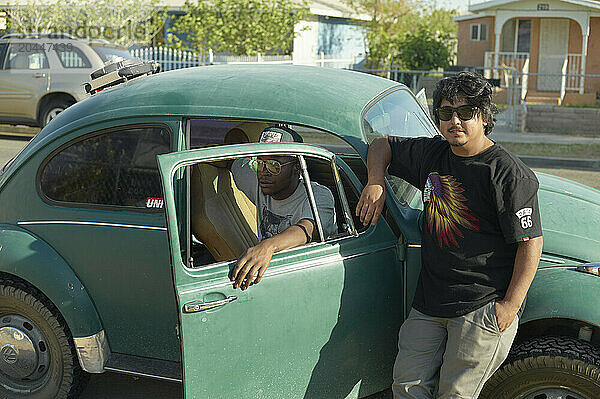 A teenaged boy leans on a vintage green car  with another teen sitting in the driver's seat; both are casually dressed on a sunny day.