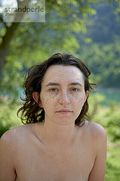 A portrait of a young woman with freckles  brown hair  and a neutral expression  standing outdoors with a blurred natural background.