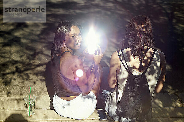 Two women sitting on steps outside  with one turning her head to smile at the camera while the other faces away. Sunlight filters through leaves  casting dappled shadows around them  and a lens flare adds a vibrant touch to the image.