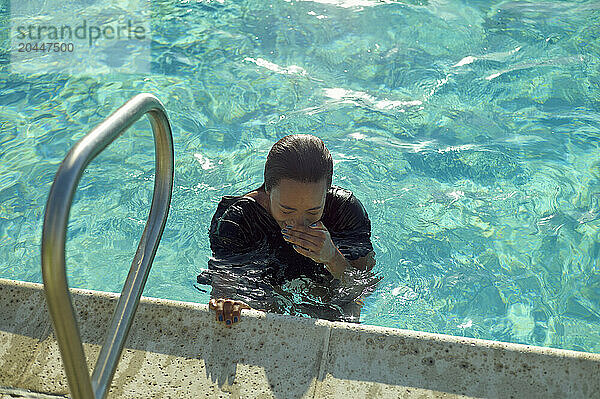 A person with wet hair is immersed in a swimming pool  holding onto the edge near a pool ladder  blowing bubbles in the water.