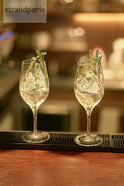 Two elegant glasses filled with clear liquid and garnished with sprigs of herbs are presented on a bar counter  with a warm  softly blurred background.