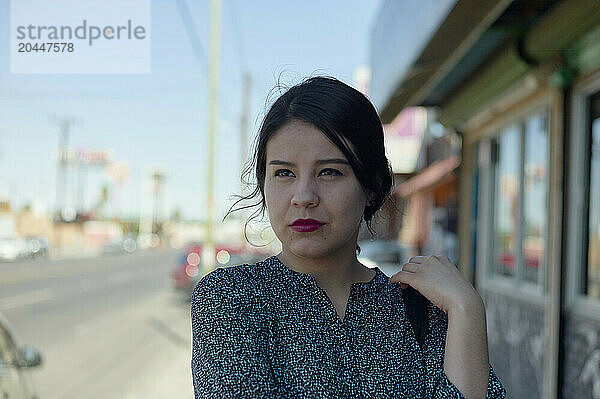 A woman stands on a sidewalk with a thoughtful expression  holding her hair  with storefronts in the blurred background.