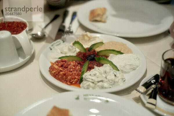 A selection of Greek dips arranged on a white plate  with cucumber slices and a black olive garnish  alongside a soft drink and table setting.