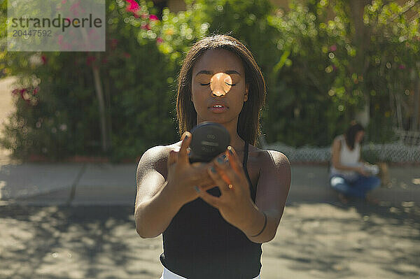 A focused young woman holds a camera lens up to her eye  taking a photo  with another person blurred in the background sitting on a bench surrounded by green foliage.