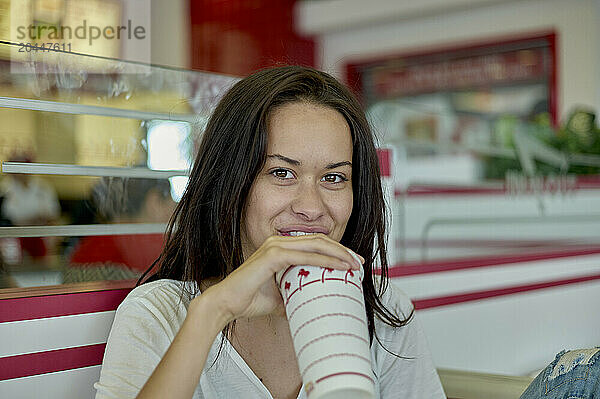 A smiling woman is casually dressed and drinks from a striped paper cup with a lid  seated in a diner with a retro aesthetic.