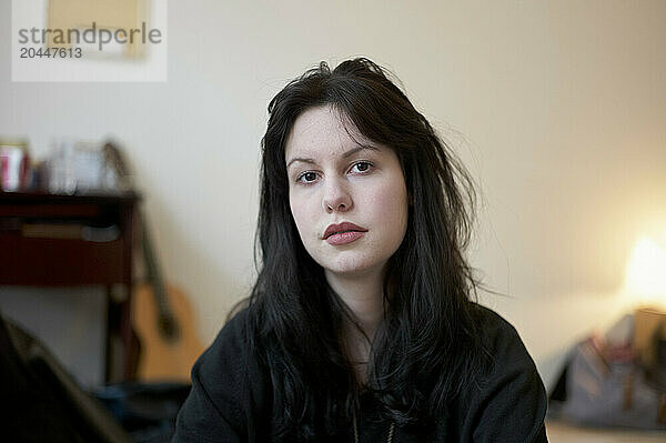 A woman with dark hair sits in a room  with some items such as pictures and a guitar blurred in the background.