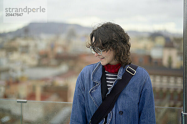 A person with curly hair smiles gently while wearing a denim jacket  patterned scarf  and glasses  with a cityscape blurred in the background.