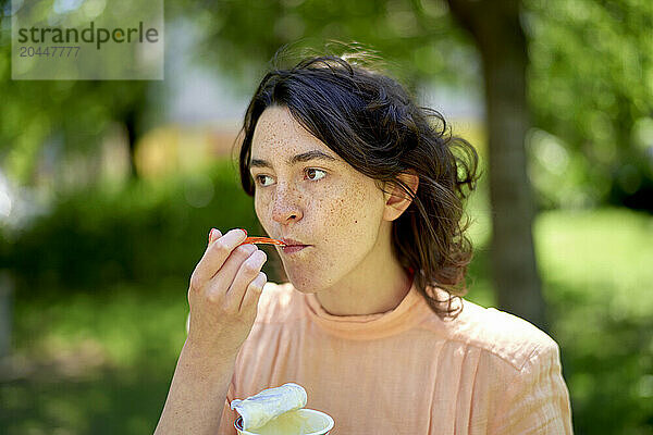 A young woman with freckles is eating with a plastic spoon outdoors  with trees and a building softly blurred in the background.