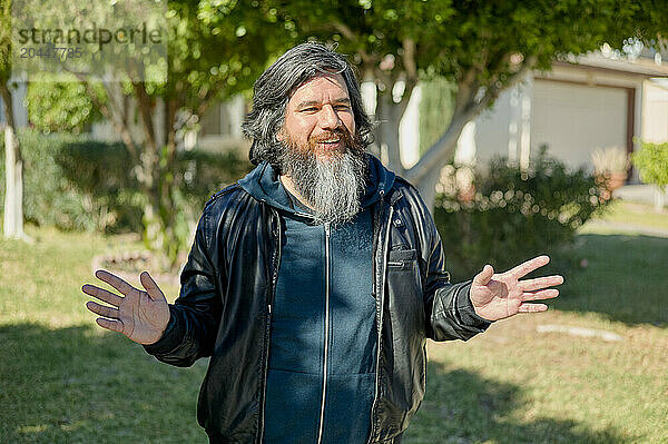 A man with a grey beard is standing outdoors  smiling and gesturing with his hands open as if he is speaking or explaining something.