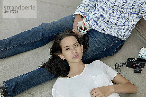 A woman is reclining on a man's lap on the ground  holding a drink and looking up at the camera  with a camera and a smartphone beside them.