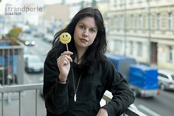 A woman stands on a balcony holding a smiley face lollipop  with a blurred city street background.