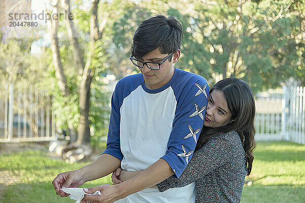 A young adult male and female are closely engaging with each other outdoors  with the female embracing the male from behind while they both look down at an object in the male's hands.