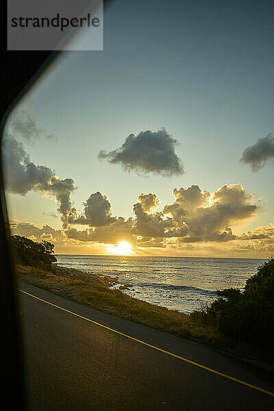 A view from a car window capturing a stunning sunset over the ocean with clouds scattered across the sky and a coastline on the left.