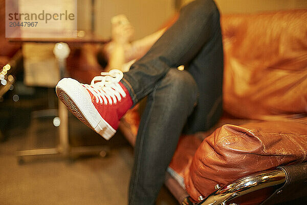 A person lounging on an orange leather couch with their legs crossed wearing red sneakers and blue jeans  with a blurred background.