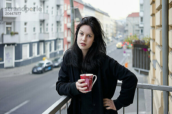 A woman stands on a balcony holding a red mug  with a street scene in the background.