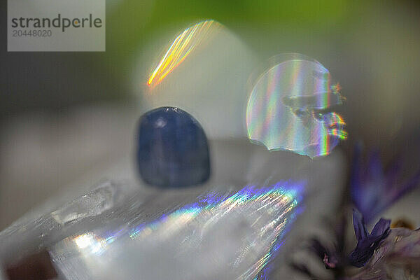 A close-up  blurred image of a crystal with a prism effect casting rainbow light refractions  accompanied by an indistinct blue object and plant material.