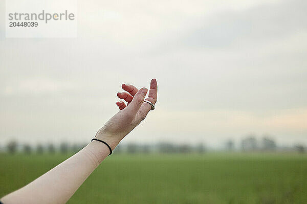 An outstretched arm reaching towards the sky with an open hand against a background of a blurred green field and a pale sky.