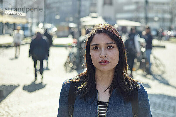 A woman stands in focus in the foreground with a blurred city street scene behind her.