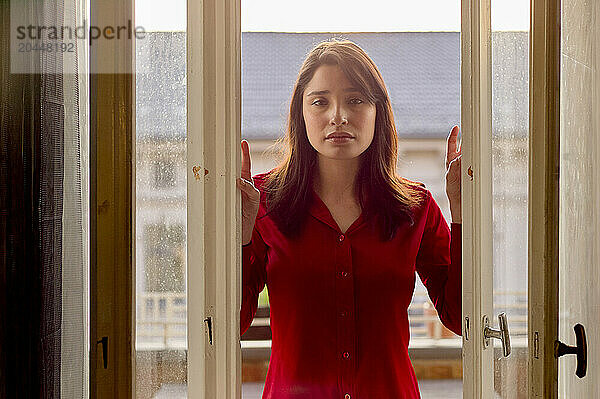 A woman in a red blouse stands between open doors  her expression contemplative as she gazes forward  with a blurred building backdrop.