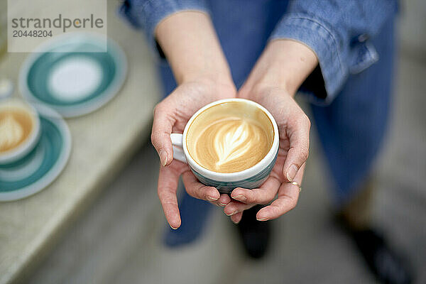 Hands cradling a ceramic cup filled with freshly brewed coffee  featuring a heart-shaped latte art  with more cups and saucers on a table in the blurred background.