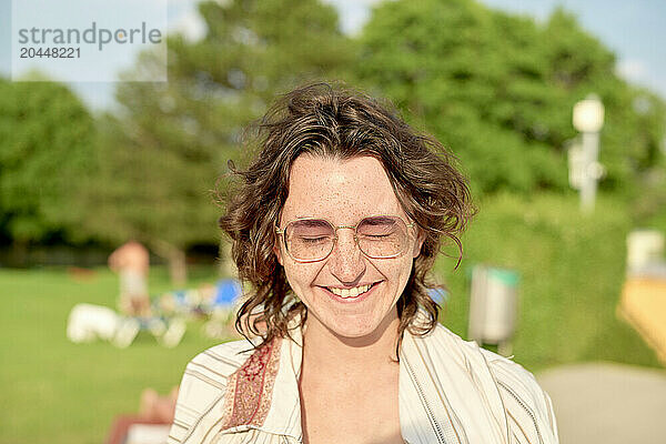 A smiling young woman with curly hair and glasses is standing outdoors on a sunny day  with blurred greenery and people in the background.