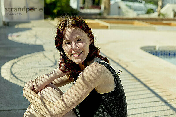 A smiling woman with red hair sitting outdoors with a swimming pool and palm trees in the background.